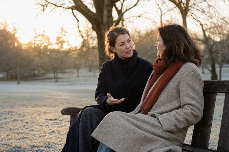 Two women in their late 40s talking on a park bench on a cold winter afternoon, one mid-sentence and the other listening