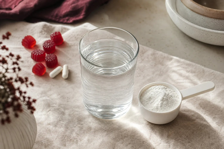 A clear glass of water, a scoop of fine white creatine powder, red gummies and two capsules on a linen counter, illustrating creatine HCl vs monohydrate and other creatine forms