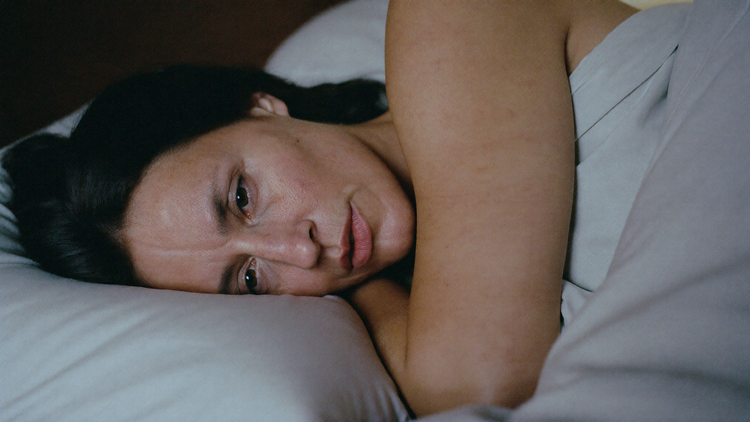 Woman in her late 40s lying awake on her side in bed at night, sheets pushed back, soft moonlight