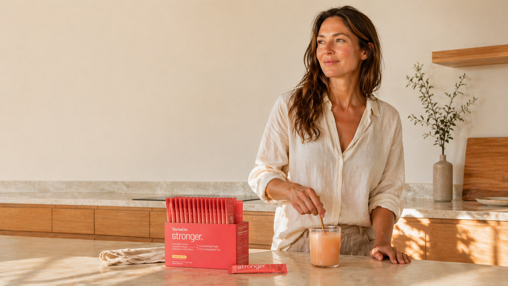 Midlife woman mixing a glass of ThriveOn Stronger in a sunlit kitchen beside the orange-mango stick pack box — daily strength support for women.
