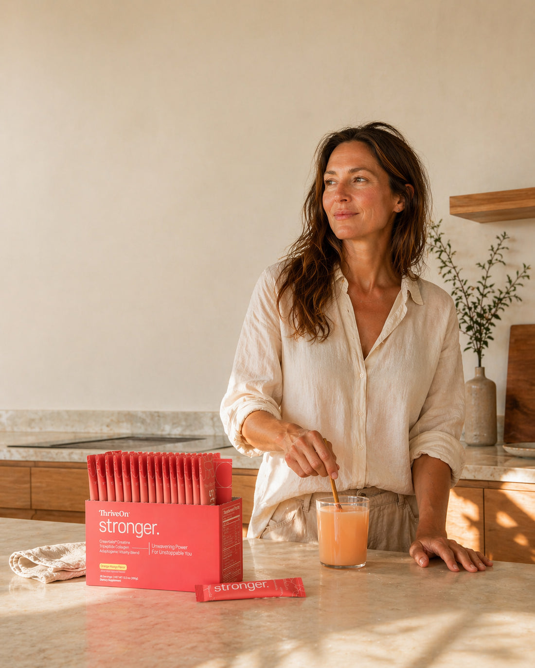 Midlife woman mixing a glass of ThriveOn Stronger in a sunlit kitchen beside the orange-mango stick pack box — daily strength support for women.